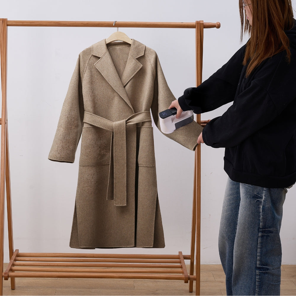 Person using fabric shaver on a beige coat on a wooden rack against a white wall