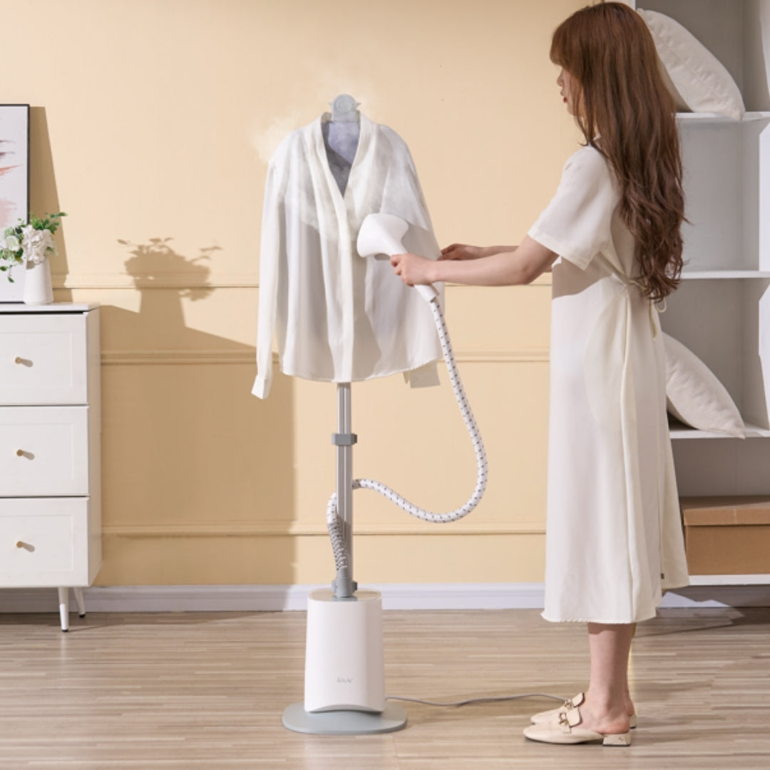Woman using a garment steamer on a white shirt in a room with a beige wall and white dresser.