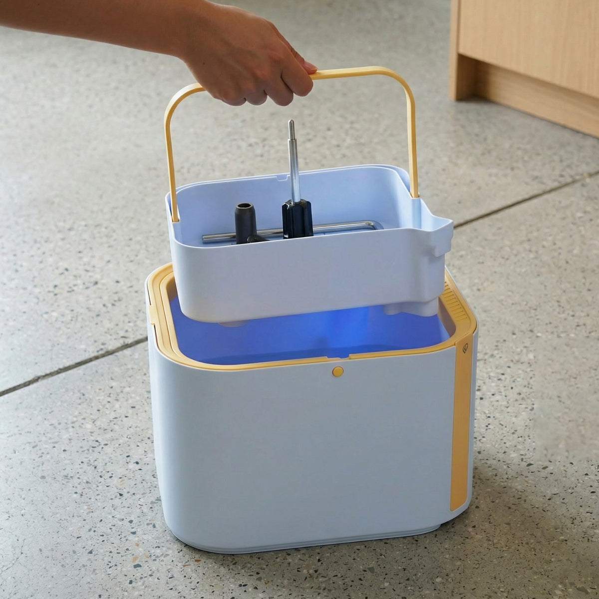 A light blue and gold plastic storage caddy with a handle, being held by a hand. The caddy has a larger base container with a blue interior and a smaller white tray on top.