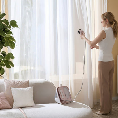 SS1500+RoseGold+Woman using a handheld steamer near a window with a white curtain in a living room.