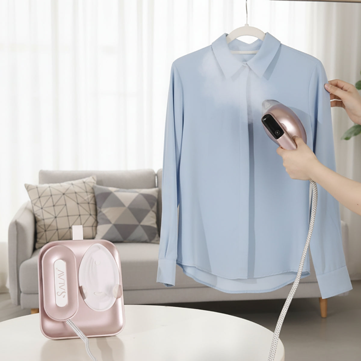 SS1500+RoseGold+Person using a handheld steamer on a light blue shirt in a living room setting.