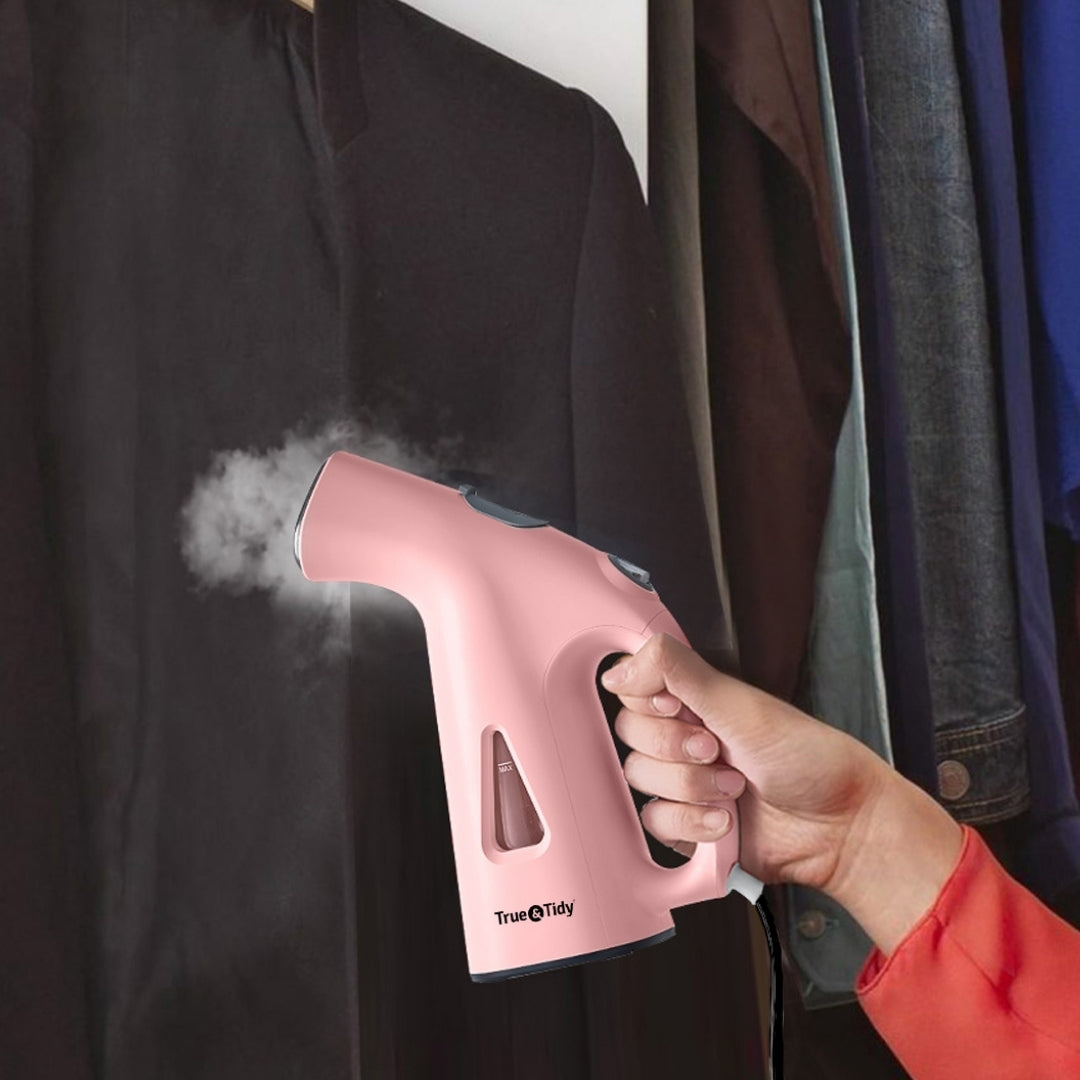 TS38 + Pink + Pink handheld steamer being used on a dark shirt with clothes hanging in the background.